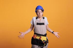 Construction worker doing i dont know sign and being clueless, acting uncertain and unsure in studio. Female contractor wearing hardhat and overalls, looking doubtful over background. photo