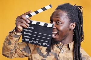 sonriente hombre participación cinematografía claqueta, posando confidente en frente de cámara en estudio terminado amarillo antecedentes. africano americano persona trabajando en película producción industria foto