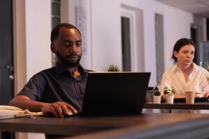 Employee working on laptop in coworking space, corporate worker sitting at workplace desk in office at night. African american man using computer in co working place with warm light photo