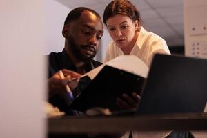 Project manager helping coworker checking report statistics, comparing data on laptop in office at night. Two employees checking marketing research results together in coworking space photo