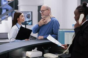 Old patient with cervical neck collar explaining fracture pain to doctor discussing health care treatment, standing at hospital registration desk. Man with medical foam waiting to attend consultation photo