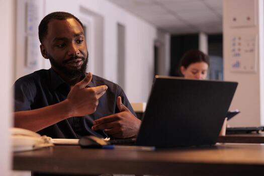 Project manager discussing company strategy plan on videoconference with coworker, corporate worker answering internet call in office at night. African american man using videocall computer software photo