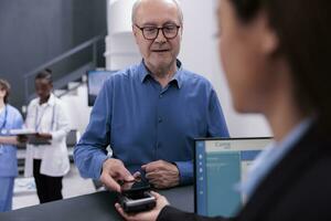 Elderly patient using phone contactless to pay checkup visit and prescription treatment at facility reception counter with receptionist. Man paying for examination appointment in waiting area lobby. photo