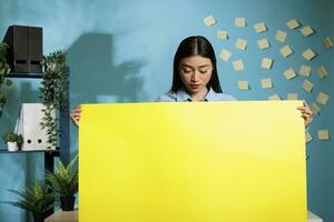 Employee of a company holding an empty banner with advertising space in a contemporary office. Startup worker standing with blank placard in hands with blue wall in background. photo