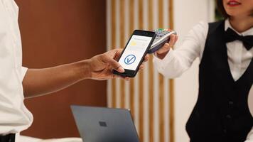 Close up of african american tourist using smartphone banking app to pay for room upgrade during check in process. Receptionist using POS payment terminal to validate credit card purchase photo