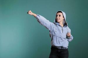 Smiling woman with hood on posing as superhero while acting like flying over isolated background in studio. Model doing justice defender gesture having power being ready to save the world, serious photo