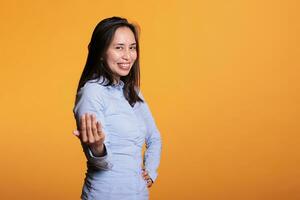 Positive cheerful filipino woman doing come to me gesture, beckoning with finger posing in studio over yellow background. Smiling model inviting you for confidential talk. Calling symbol photo