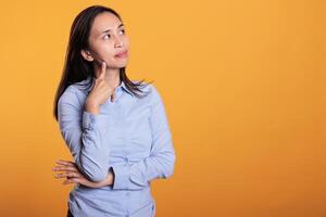 Doubtful asian woman thinking with finger on chin in front of camera in studio. Pensive model feeling confused standing over yellow background. Pensive adult showing contemplative expression photo