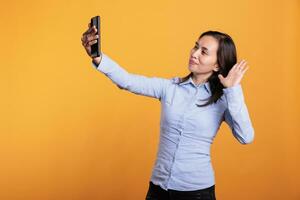 Filipino cheerful woman greeting remote friend during mobile phone videocall meeting, enjoying conversation in studio over yellow background. Attractive adult having fun during smartphone call photo