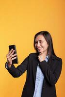 Cheerful asian woman talking with remote friend during videocall meeting conference, having remote discussion during shoot break in studio. Adult in formal suit standing over yellow background photo