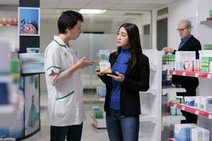 asiático mujer participación vitamina paquete y escuchando a farmacéutico explicando ascórbico ácido Dosis instrucción. farmacia cliente comprando comida suplemento, hablando con farmacia asistente en pasillo foto