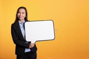 Filipino woman holding isolated speech bubble in studio, using blank copyspace cardboard to create new advertisement. Cheerful young adult showing carton ad on camera over yellow background photo