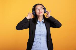 filipino alegre mujer vistiendo auriculares escuchando música, teniendo divertido durante descanso en estudio terminado amarillo antecedentes. sonriente modelo disfrutando en línea podcast utilizando auriculares. entretenimiento concepto foto