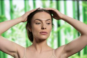 Attractive woman touching head, using organic cosmetics on hair roots. Beautiful young model massaging scalp, doing haircare routine and posing with neutral facial expression photo