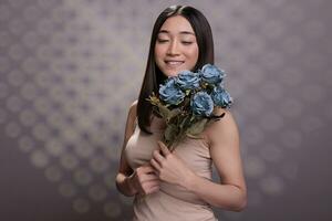 sonriente asiático mujer participación presentado rosas ramo de flores en hombro y mirando en raro azul flor brotes joven hermosa dama posando con único creativo flores regalo en estudio foto