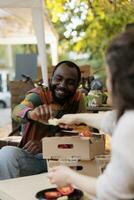 Happy young African man customer tasting organic fruits and vegetables at farmers market, smiling black guy sitting at table with local vendor discovering apple varieties while visiting food fair photo