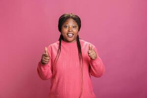 sonriente africano americano joven adulto haciendo pulgares arriba firmar mientras posando en estudio terminado rosado antecedentes. contento alegre mujer con alegre facial expresión demostración aprobación gesto, persona haciendo me gusta foto