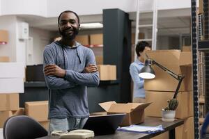 Smiling warehouse employee standing with arm crossed working at clients orders while preparing cardboard boxes in storehouse delivery department. In background woman checking packages shipping details photo