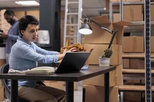 Supervisor looking at trendy blouse putting client order in cardboard box after checking personal details on laptop computer, preparing for shipping Team working in warehouse delivery department photo