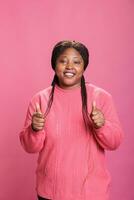 Smiling african american model showing thumbs up sign standing over pink background in studio. Happy cheerful woman with joyful facial expression giving approval gesture, person doing like sign photo