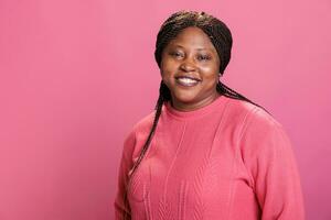 retrato de africano americano modelo sonriente a cámara mientras posando contento en estudio con rosado fondo, teniendo positivo expresión. confidente mujer con elegante peinado y rosado sudadera foto