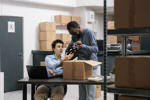 Storehouse supervisor sitting at desk looking at cargo stock while preparing client package for delivery, worker holding helmet order discussing shipping detalies. Team working in warehouse photo