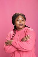 retrato de mujer sonriente con confianza mientras posando en estudio teniendo alegre expresión disfrutando disparar tiempo. africano americano modelo con elegante peinado y rosado sudadera mirando a cámara foto