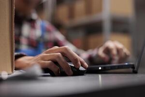 Postal warehouse employee checking delivery customer satisfaction on laptop. Post office storehouse worker managing shipping operations while using computer mouse close up photo