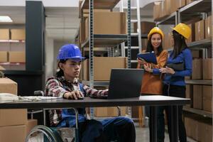 Asian warehouse employee in wheelchair doing goods inventory optimization on laptop. Postal service shipment operator working on computer at desk in disability friendly storehouse photo