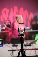 Woman with pink hair having fun while recording video with her performing techno music at professional mixer console, filming process with phone. Artist dancing while mixing song at night in club photo