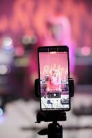 Cheerful artist with pink hair standing at dj table mixing song with turntables in front of camera while recording video with phone. Smiling woman doing performance at nightclub with audio equipment photo