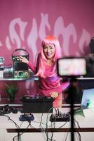Cheerful dj mixing electronic music at mixer console having fun while performing song at night in club. Artist with pink hair recording performace with camera posting video online for subscribers photo