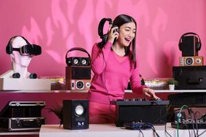 Asian musician with headphones playing electronic song at mixer console, standing at dj table having fun with fans at night in club. Artist performing techno music in studio over pink background photo