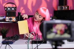 Asian influencer holding notebook advertising product in front of camera while recording school supplies review using broadcast equipment. Content creator with pink hair live streaming photo