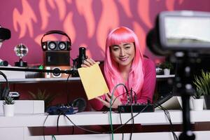 Influencer with pink hair advertising notebook in front of camera recording vlog while presenting school supplies review using professional vlogging equipment. Asian creator filming podcast photo