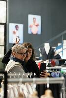 sonriente mujer tomando selfie con móvil teléfono en sala de exposición, teniendo divertido durante compras sesión. alegre clientes comprobación formal vestir marcas para comercial actividad, comprando de moda ropa foto