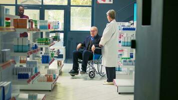 Pharmacist giving cardiology pills bottle to senior wheelchair user, man with chronic disability asking for support. Client with impairment receiving pharmaceutics treatment in pharmacy. photo