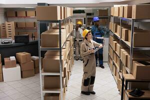 Delivery service worker searching customer parcel in warehouse and holding clipboard. Diverse storehouse employees wearing uniform overalls supervising goods in factory storehouse photo