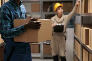 African american man lifting heavy parcel in distribution warehouse, carrying carboard box to stockroom shelf. Storehouse loader holding carton and working in logistic department goods storage photo