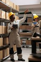 Woman storehouse operator working with digital tablet in warehouse and taking parcel from shelf. African american delivery service storage employee pointing at cardboard boxes photo