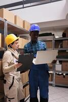Logistic department warehouse workers monitoring freight distribution on laptop and discussing inventory management system. Two storehouse employees supervising stock arrangement photo