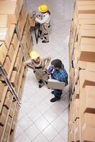 Warehouse supervisors analyzing stock management system on laptop and packing parcels with adhesive tape. Shipping company employees preparing cardboard boxes for sending and chatting top view photo