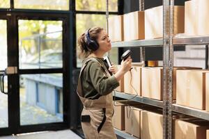 Small business employee scanning carton boxes barcode using store scanner, listening music during warehouse inventory. Worker looking at storehouse shelves, preparing clients orders for delivery photo