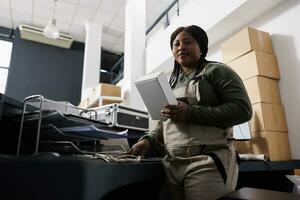 Portrait of african american worker holding white box, preparing customers orders before shipping packages. Stockroom employee wearing industrial overall checking merchandise checklist photo