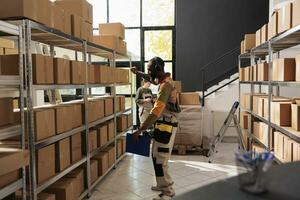 Warehouse supervisor listening music and dancing during inventory, checking cardboard boxes details before shipping to customers. African american worker with protective overall working in storage photo