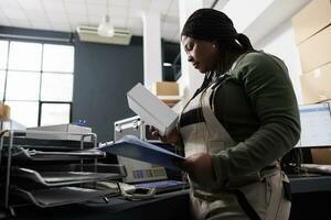 African american worker looking at white cardboard box, preparing customers orders at counter desk in storehouse. Stockroom employee wearing industrial overall during goods inventory photo