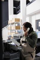 African american worker looking at white cardboard box while talking with remote client, discussing online order using landline phone. Stockroom supervisor working at goods inventory photo