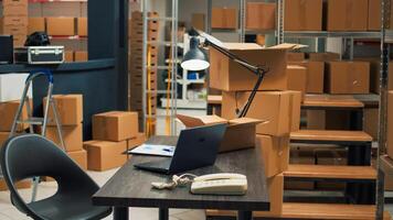 Empty desk room in storage warehouse area filled with carton boxes, office with laptop and inventory or logistics notes. Storespace with stacks of merchandise in cardboard packages. photo