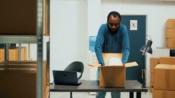 Male warehouse worker arranging supplies in packages, using laptop to plan goods inventory and merchandise logistics. Entrepreneur putting products in cardboard boxes, stock management. photo