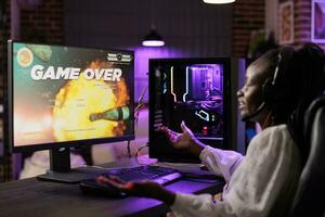 Gamer feeling irked after losing singleplayer action videogame mission, being outsmarted by enemies. African american woman confused after seeing game over screen on gaming computer photo